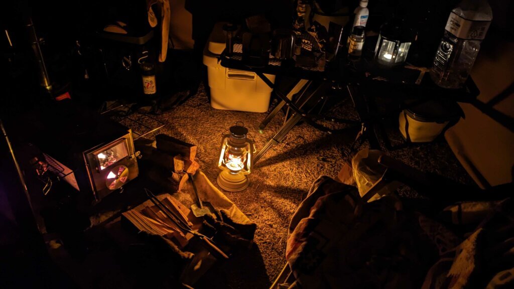 夜のテント内の様子。写真の中央ではランタンが灯っている。写真左側にはキャンプ用薪ストーブ、写真右上にはキャンドルランタンが灯っている。
